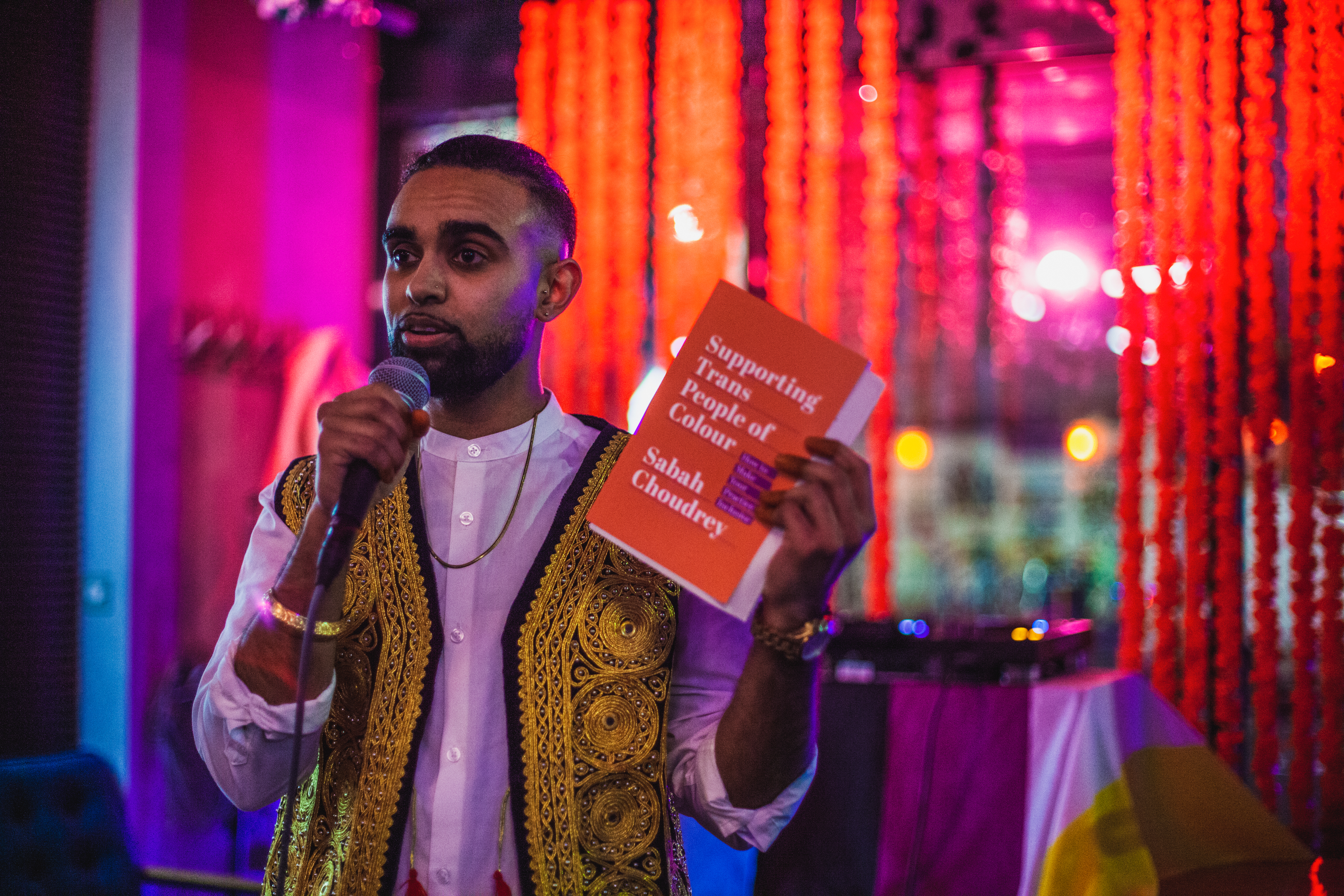 Sabah is holding a microphone in one and and holding their book, Supporting Trans People of Colour in the other hand. They have their hair tied back in a bun, wearing a white shirt, with a gold and black Pakistani style waistcoat.