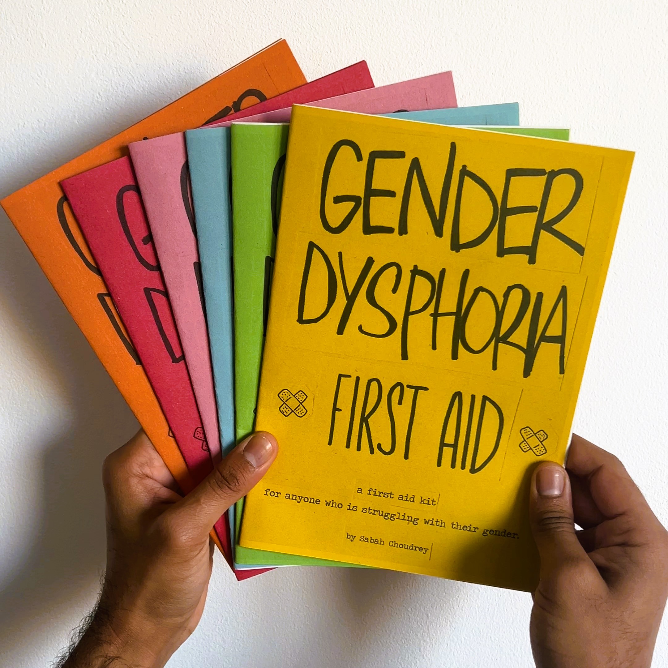 two brown skinned hands are holding 6 'gender dysphoria' zines on a white background. each zine is a different colour - orange, red, pink, blue, green and yellow. 